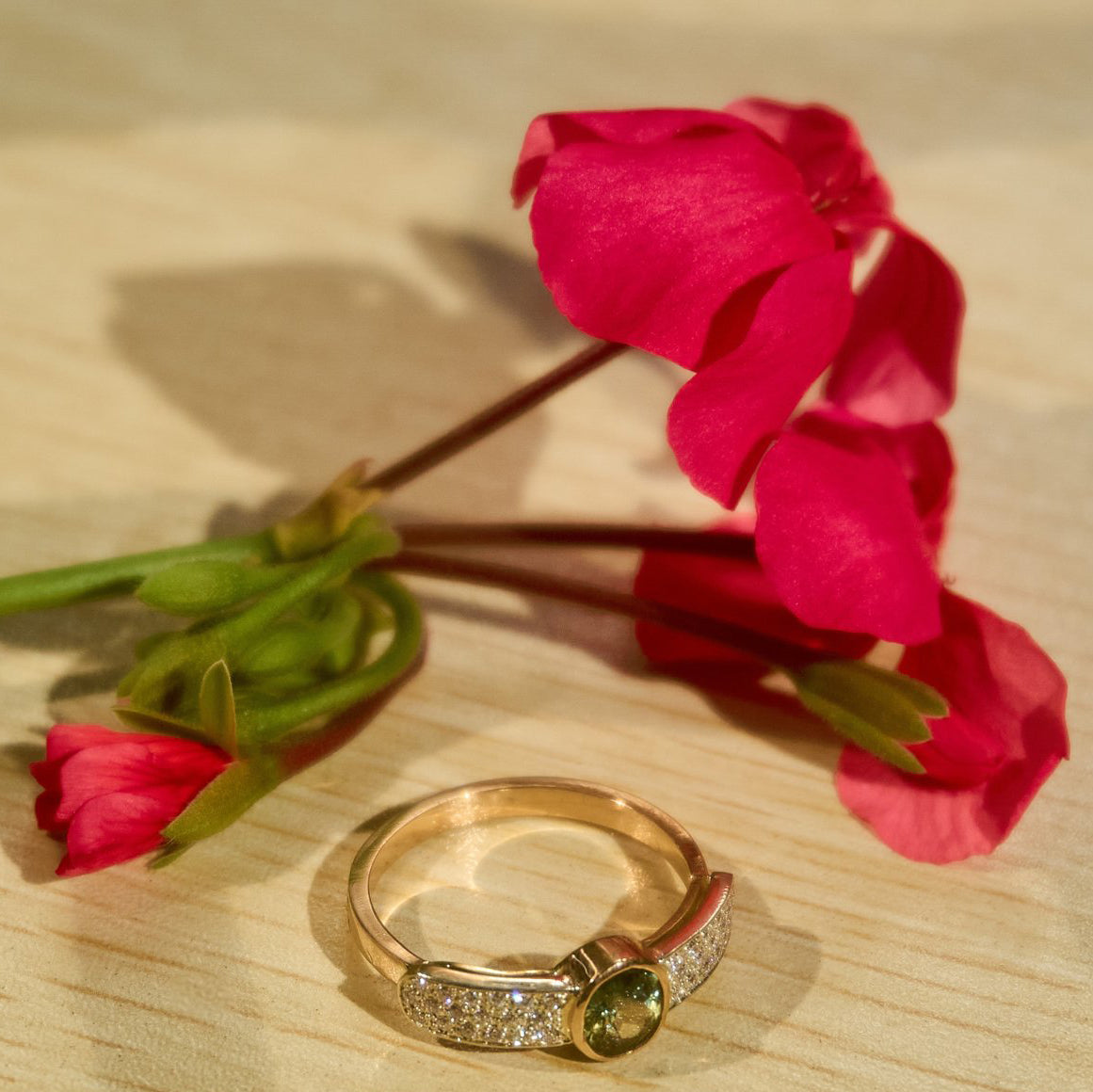 18k gold and diamond ring with green gemstone beside red flowers on wood surface