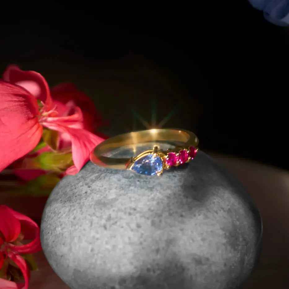 18k gold ring with pear-shaped sapphire and rubies, luxury jewelry on stone with pink flowers